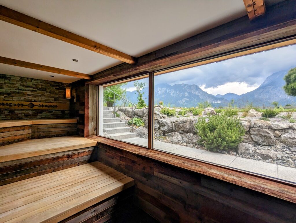 A cozy, wooden sauna interior with tiered seating is shown, featuring a large window with a view of the rocky terrain of the bavarian alps and lush greenery outside. In the background, the german neuschwanstein castle looms in the majestic mountains are partially obscured by clouds, creating a serene and natural backdrop.