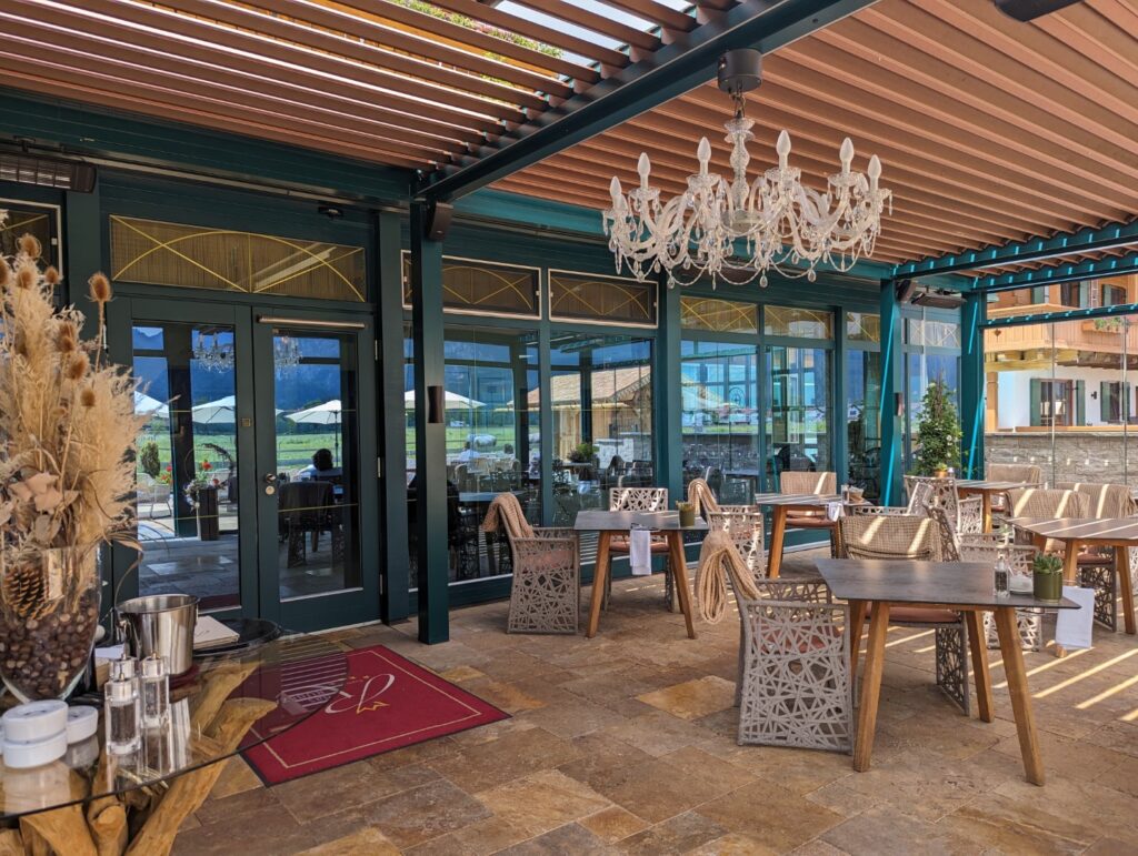 An elegant outdoor dining area at Hotel Das Rubezahl features wooden tables and chairs with intricate designs, a large chandelier, and a view of the surroundings through glass doors. The space is shaded by a pergola and decorated with dried plants and lanterns.