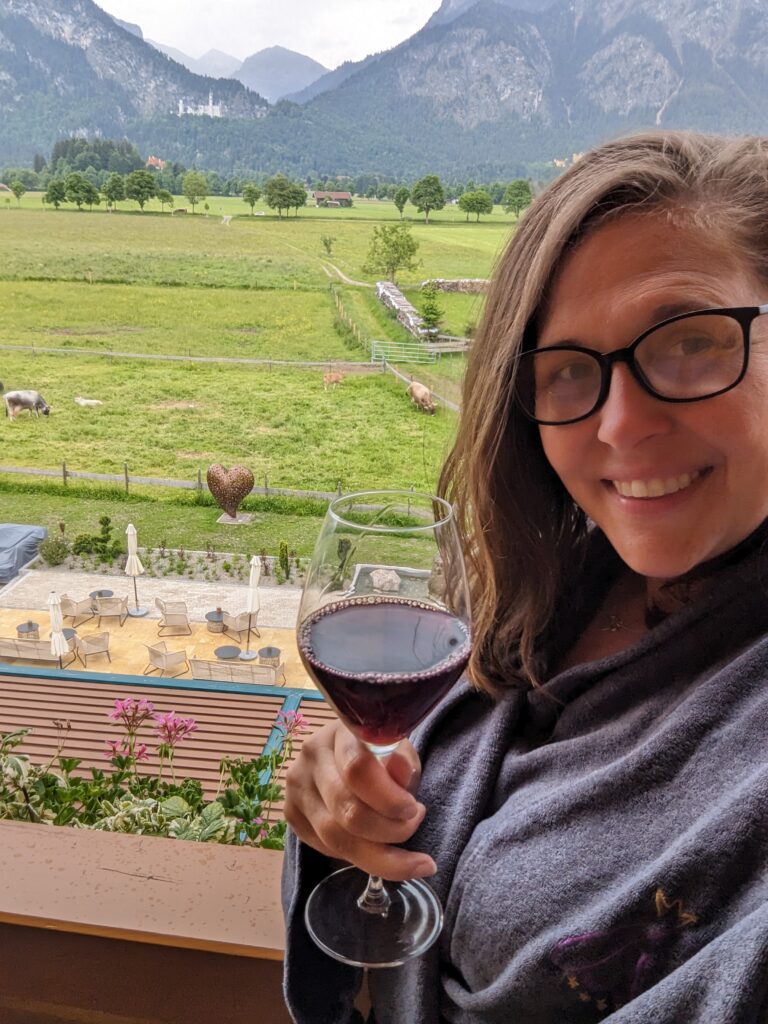 A woman wearing glasses and a dark sauna robe smiles at the camera while holding a glass of red wine. She is on a balcony overlooking a scenic landscape with green fields, distant trees, mountains, and neuschwanstein castle in the background.
