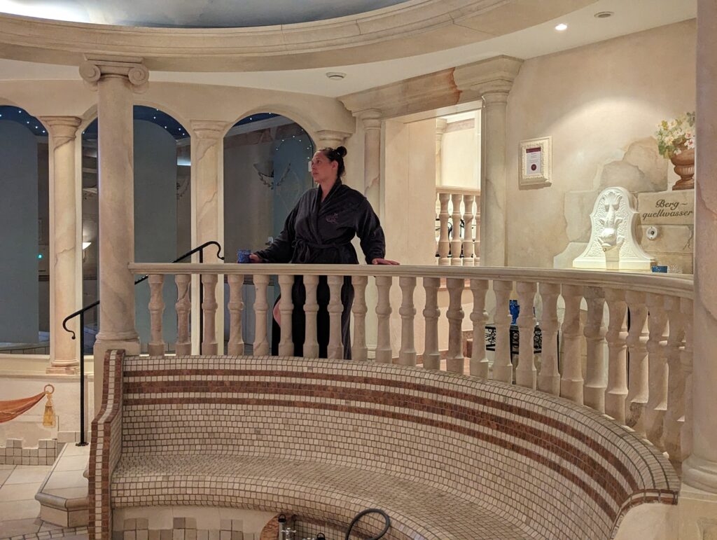 A person in a dark robe stands on a balcony inside an elegantly decorated European spa. The space features arched doorways, a sculpted bust, and intricate tile work on the walls and floors.