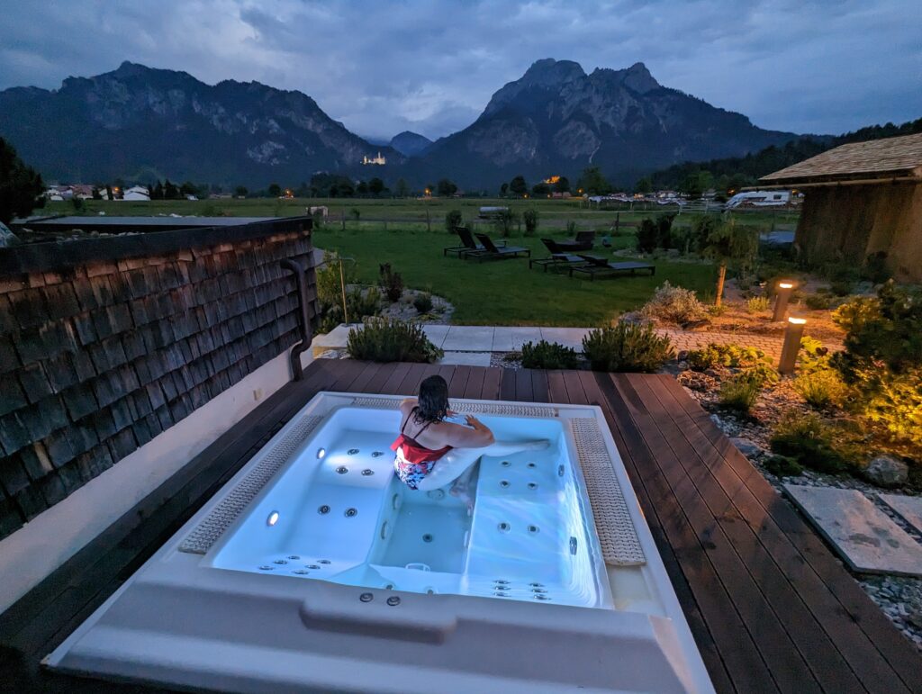 A person relaxes in an outdoor hot tub at dusk as she gazes out to the Neuschwanstein castle in Germany in the distance, with a stunning view of mountains in the background. The area is softly lit, with surrounding greenery and a wooden structure nearby.