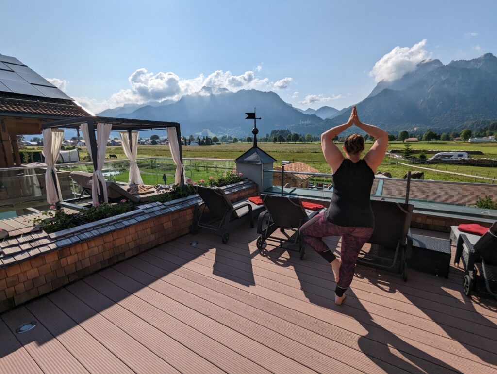 A person practices yoga on a sunlit deck with a beautiful view of green fields and distant mountains and the german neuschwanstein castle under a blue sky. The deck features lounge chairs and a canopy-covered relaxation area, creating a tranquil outdoor space.