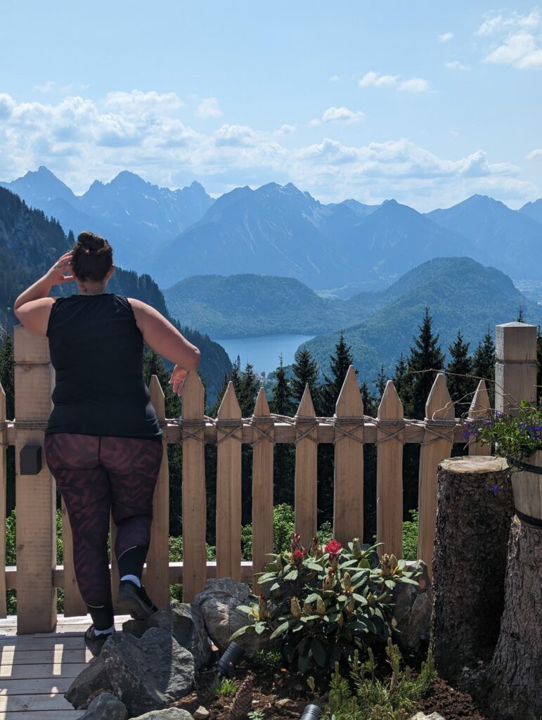 A woman in a black tank top and patterned leggings stands on a terrace, gazing at a breathtaking view of the bavarian alp mountains, lake, and neuschwanstein castle. She is leaning against a wooden fence, surrounded by some plants and flowers.